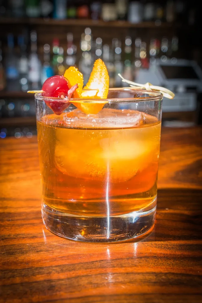 Amber Old Fashioned cocktail with spherical ice, orange peel, cherries on a bar.