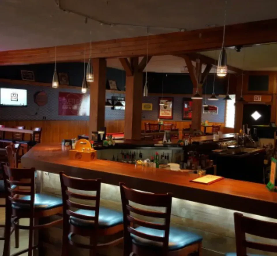 Empty, rustic bar interior with wooden counter, beams, and warm lighting.
