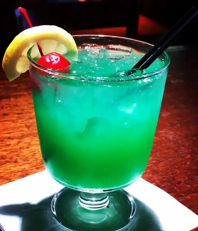 Teal-green cocktail with ice, cherry, lemon, and straws.