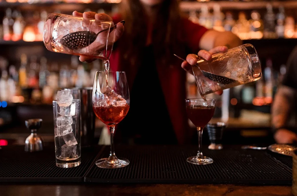 Bartender's hands pouring two cocktails into three ice-filled glasses.