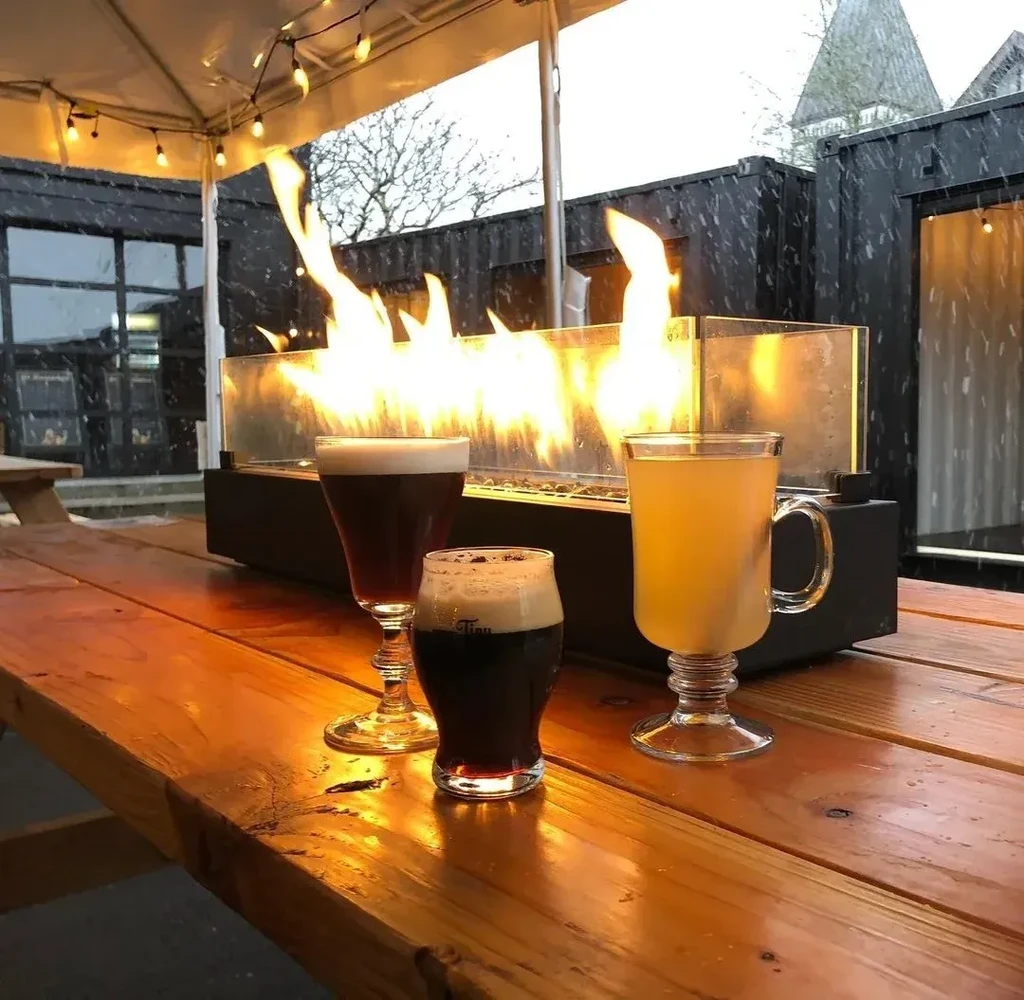 Beverages on a rustic table by a fire pit as snow falls.