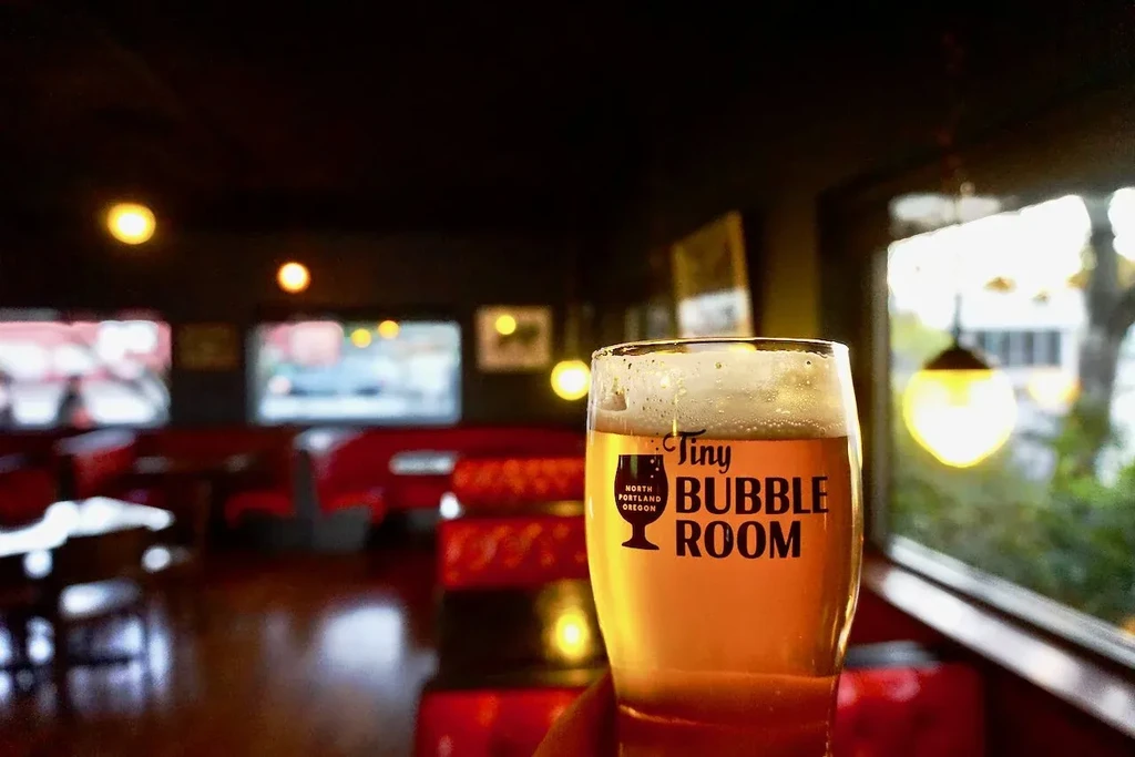 Hand holding "Tiny Bubble Room" beer glass inside a dimly lit pub.