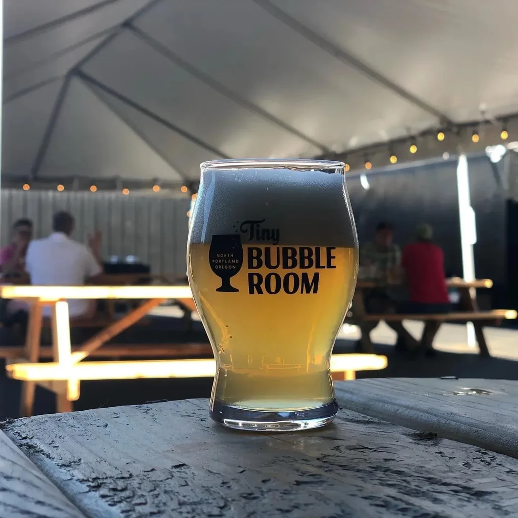 Frothy amber beer glass with Tiny Bubble Room logo outdoors.