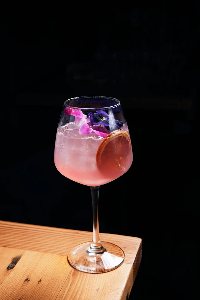 Pink cocktail in stemmed glass with flowers and dried citrus on wood.