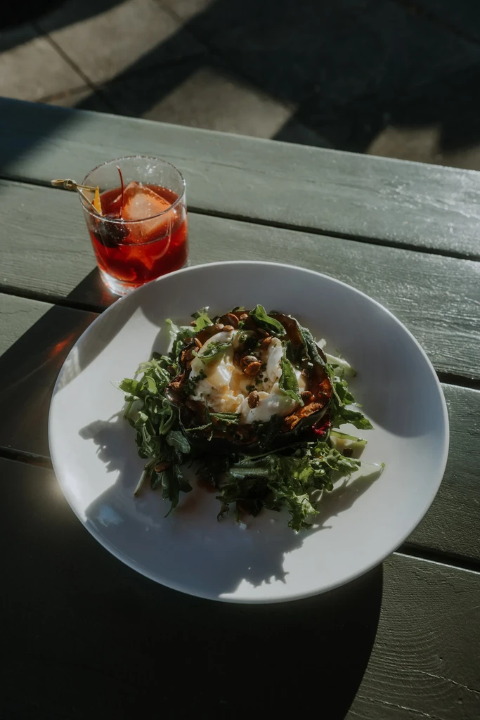 Overhead: Roasted vegetables, cheese, greens, red cocktail on green table, sunlight.