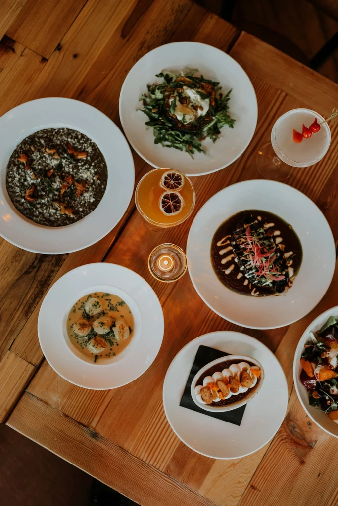 Overhead: diverse dishes, cocktails, and a lit candle on a wooden table.