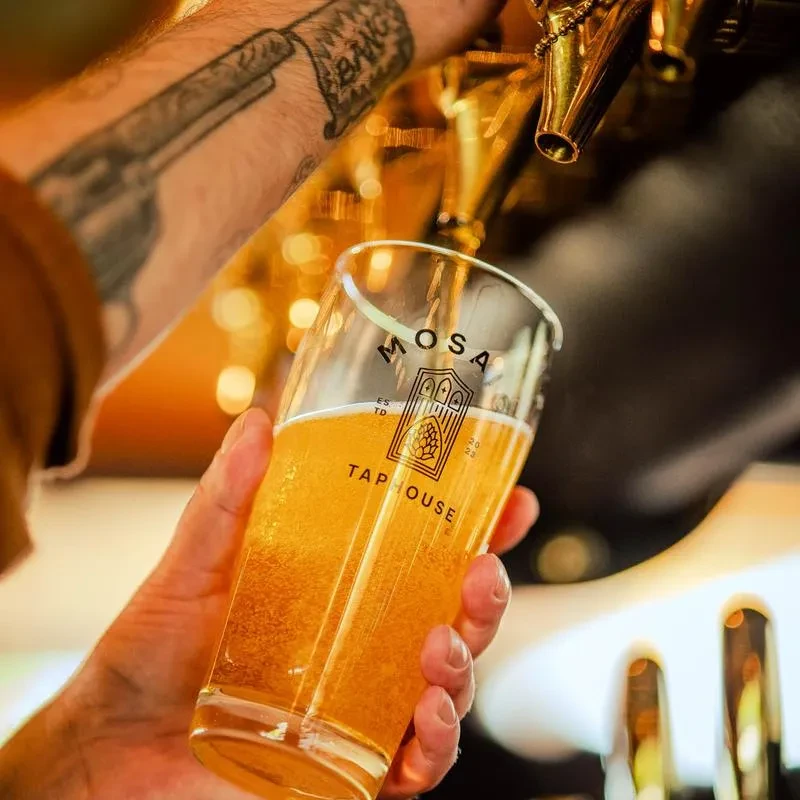 Tattooed hand holds Mosa Tap House glass, beer pouring from tap.