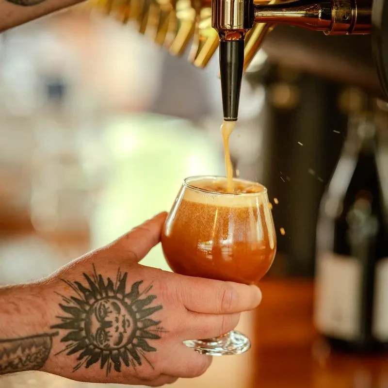 Tattooed hand holds glass as amber beer pours from golden tap.