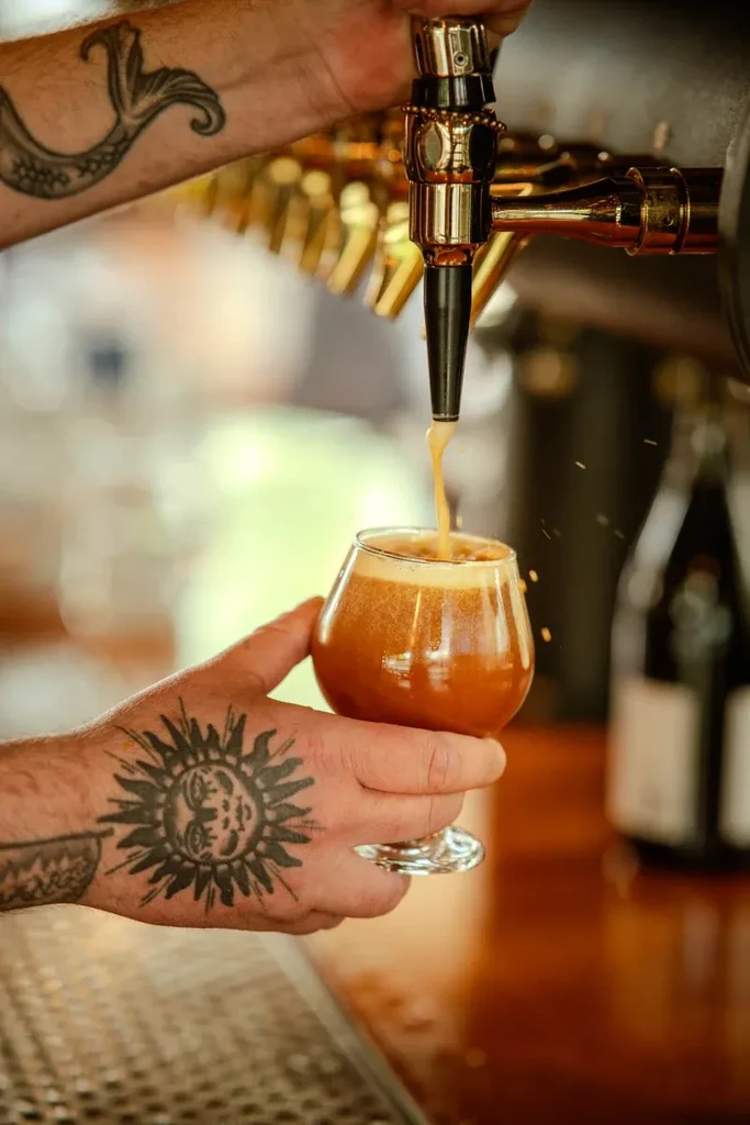 Hands pouring amber beer from a tap, with sun and mermaid tattoos.