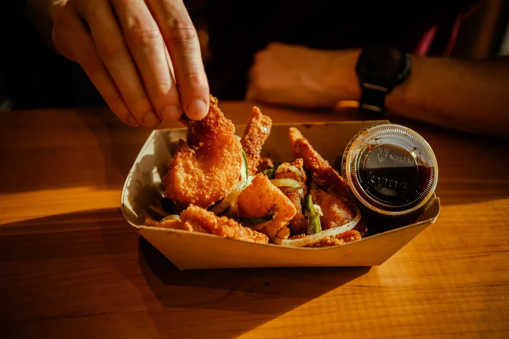 Hand picking golden-brown fried food from tray with onions, peppers, and sauce.
