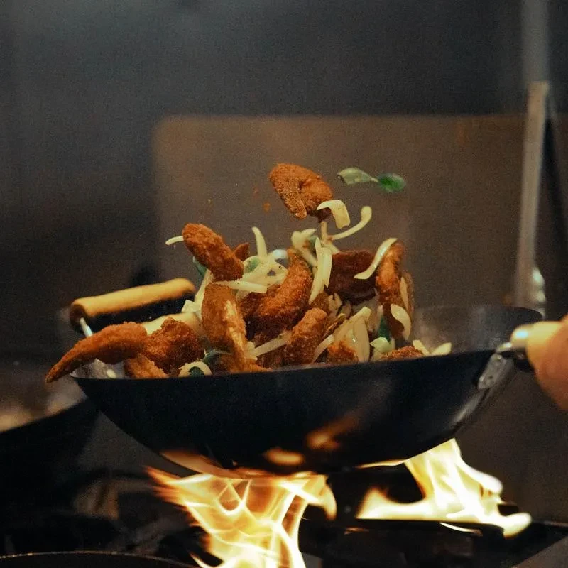 Breaded food and vegetables tossed from wok over fiery burner.