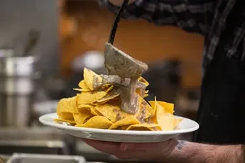 A person ladles thick sauce over a large plate of nachos.