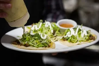 Hand adds yellow sauce to three tacos with lettuce and cream.