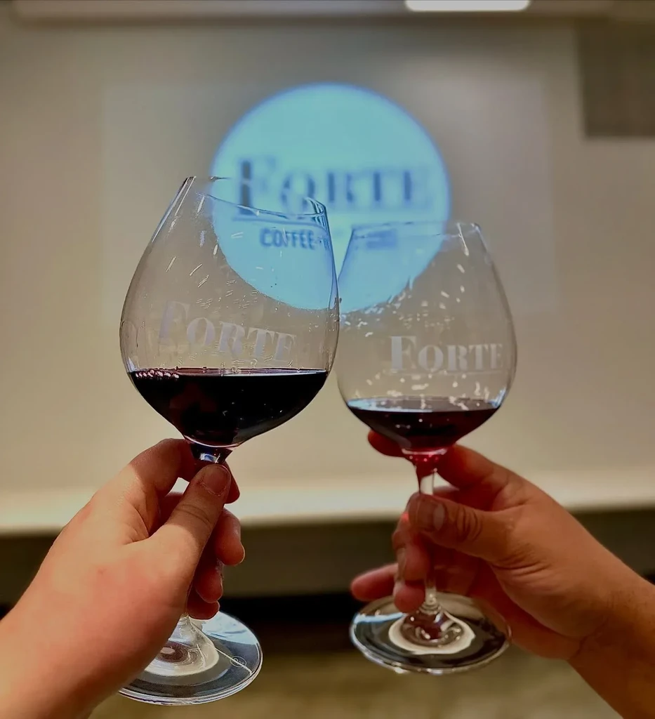 Hands toasting red wine glasses etched with "FORTE," background sign "FORTE COFFEE."