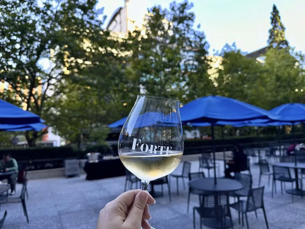 Hand holds "FORTE" wine glass on a patio with blue umbrellas.