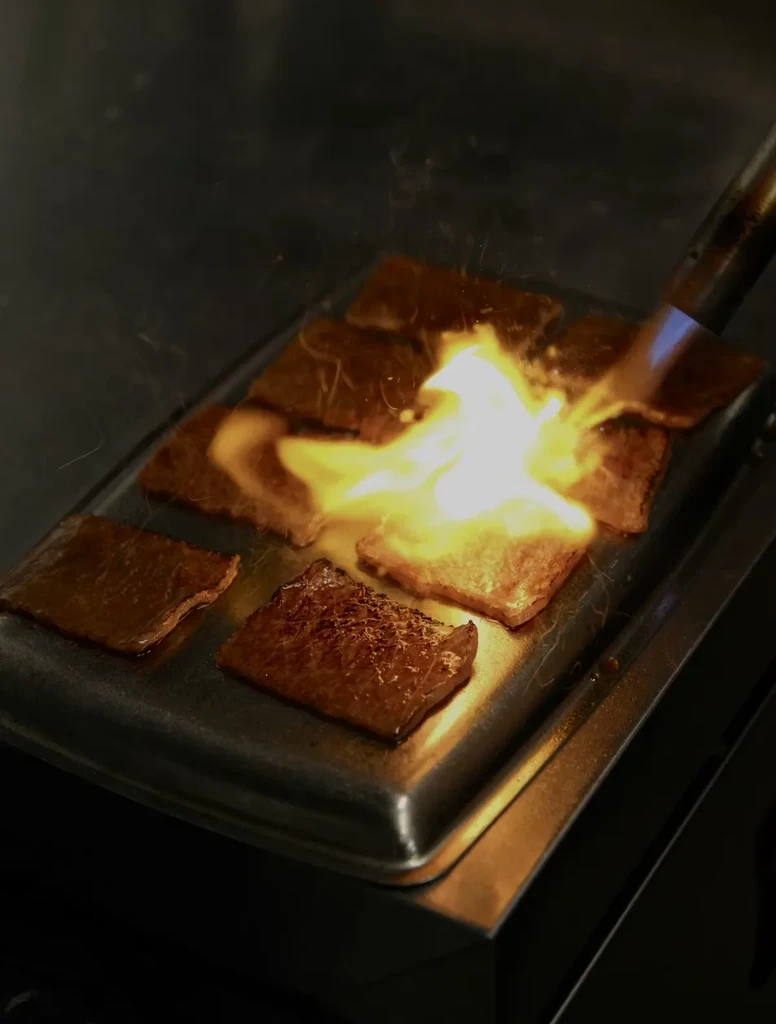 Close-up of protein cooking on griddle, ignited by a culinary torch.