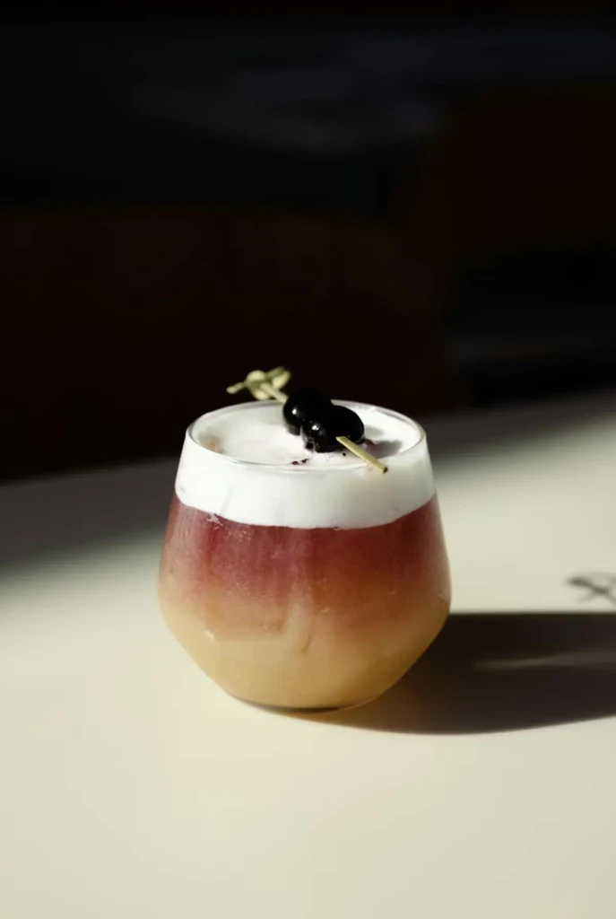 Layered cocktail: white foam, purple liquid, amber bottom, and berry garnish.