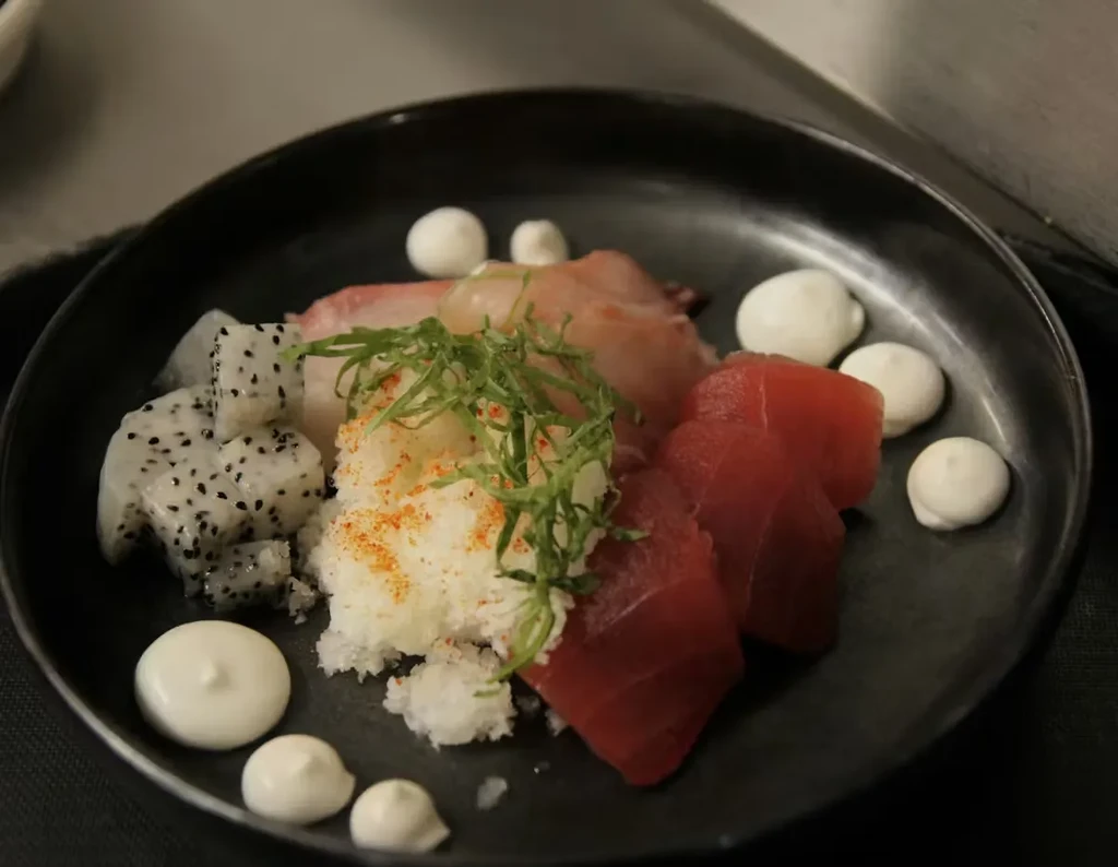 Artfully plated sashimi with red fish, dragon fruit, herbs, dark bowl.