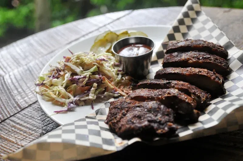 BBQ ribs on checkered paper with coleslaw, sauce, and pickles.