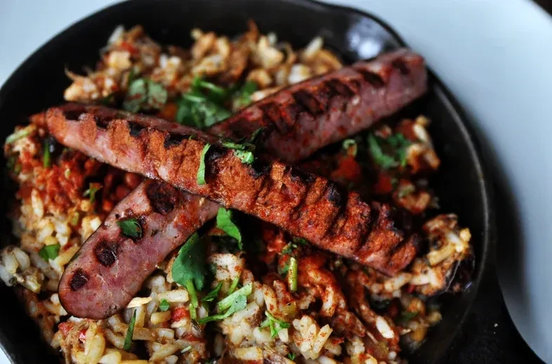 Overhead view of grilled sausages on herbed rice in a dark pan.