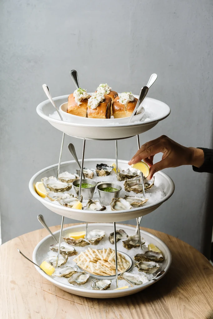 Three-tiered stand of oysters, crab rolls, crackers, and hand squeezing lemon.