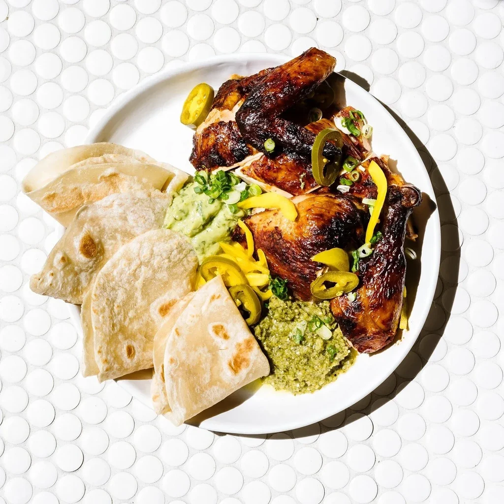 Roasted chicken, two green dips, tortillas, and peppers on a white platter.