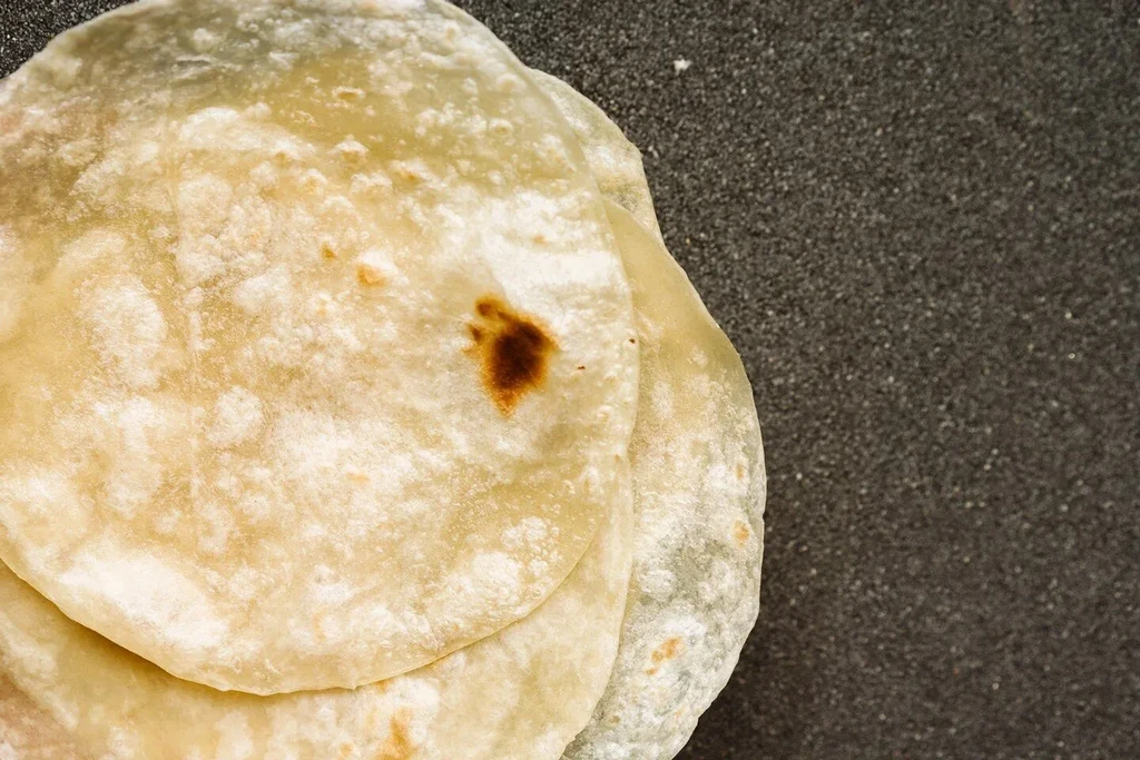 Stack of pale, freshly made tortillas with browning on a dark griddle.