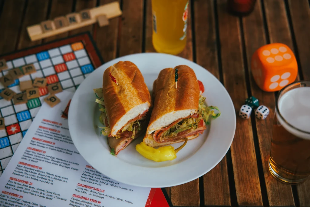 Sub sandwich, Scrabble, dice, and beer on a wooden table.