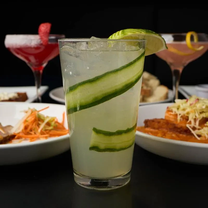 Tall cocktail with spiraled cucumber and lime; blurred drinks and food.