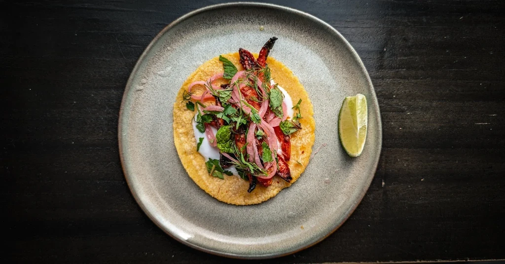 Overhead: plated corn taco with peppers, herbs, sauce, and lime.