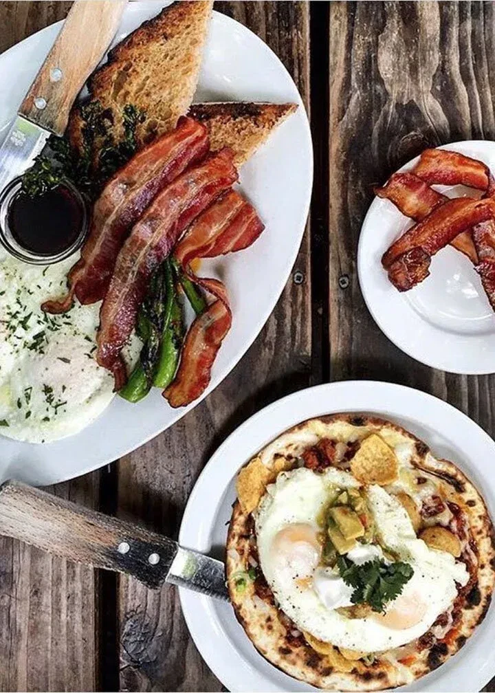Plates of bacon, eggs, toast, and flatbread on a rustic wooden table.