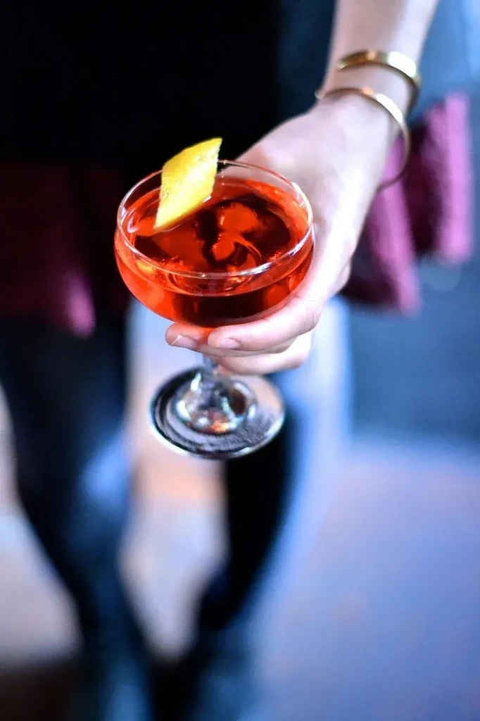 Hand with gold bangles holding red cocktail with lemon twist.