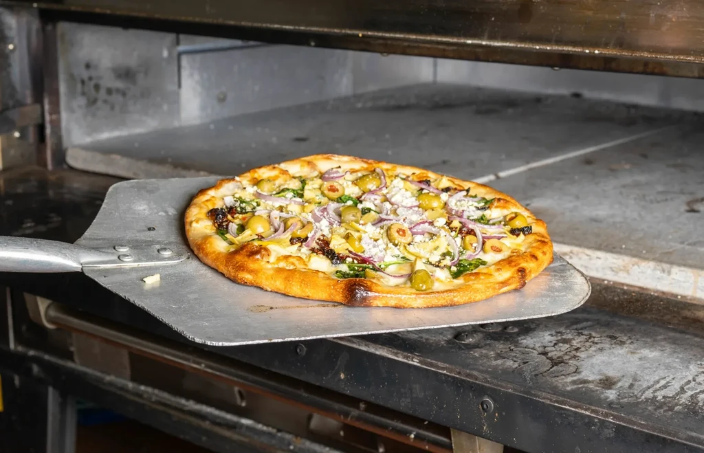 Pizza with olives, feta, onion, and tomatoes removed from a professional oven.