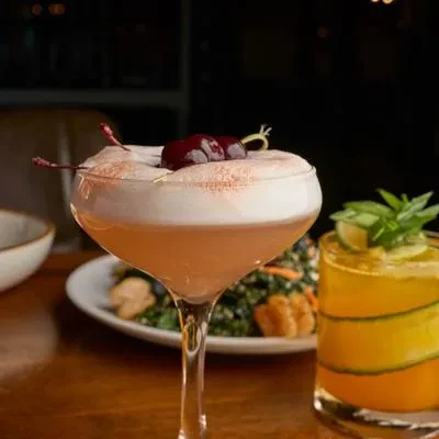 Two cocktails on a table: frothy pink, and amber with cucumber.