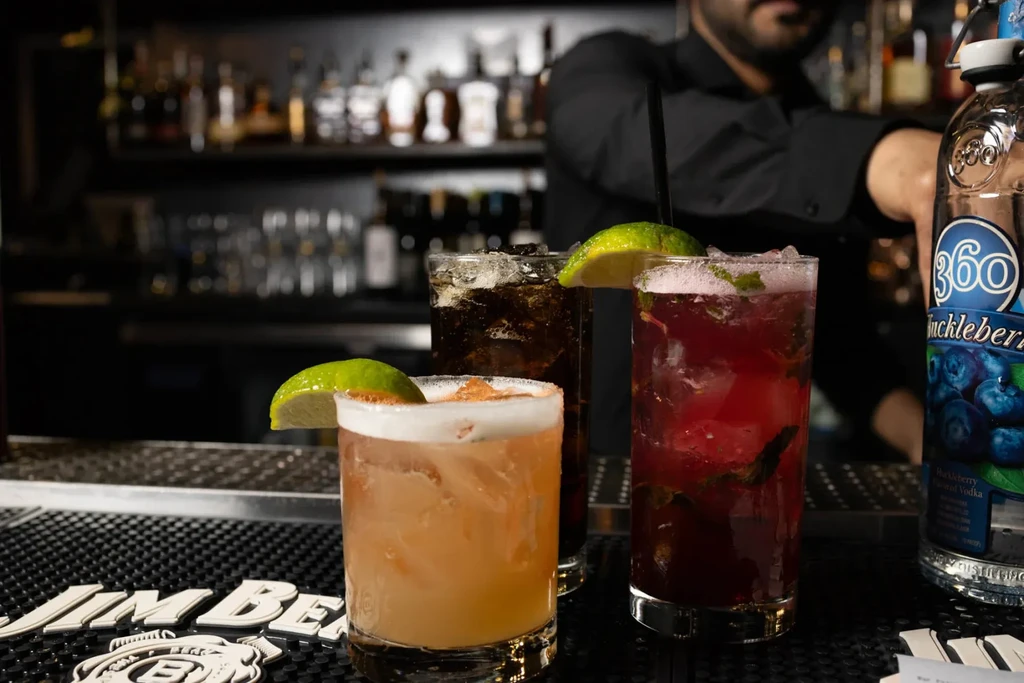 Three colorful cocktails on a bar mat, blurred bartender visible.