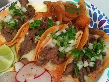 Four garnished tacos with radishes, lime, and peppers on a paper plate.