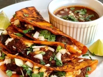 Plate with three birria tacos, consommé, cilantro, onions, and lime.