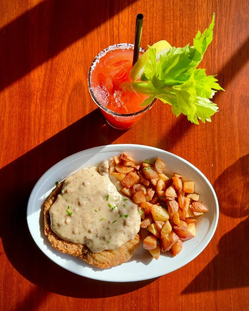 Overhead: Chicken fried steak, potatoes, Bloody Mary on a sunlit wooden table.