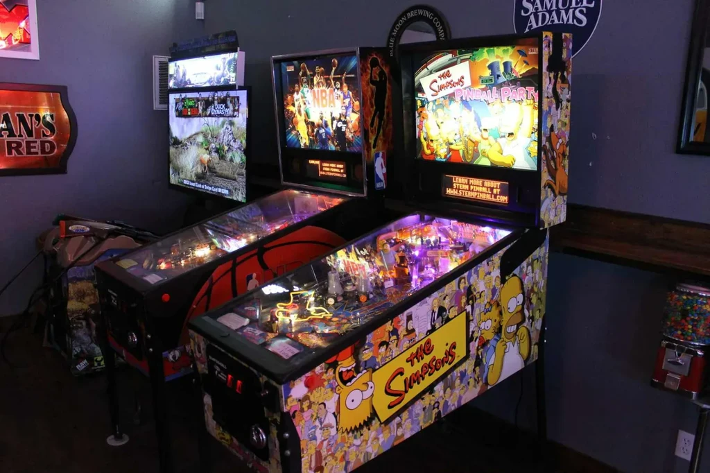Three themed arcade and pinball machines brightly lit in a bar.
