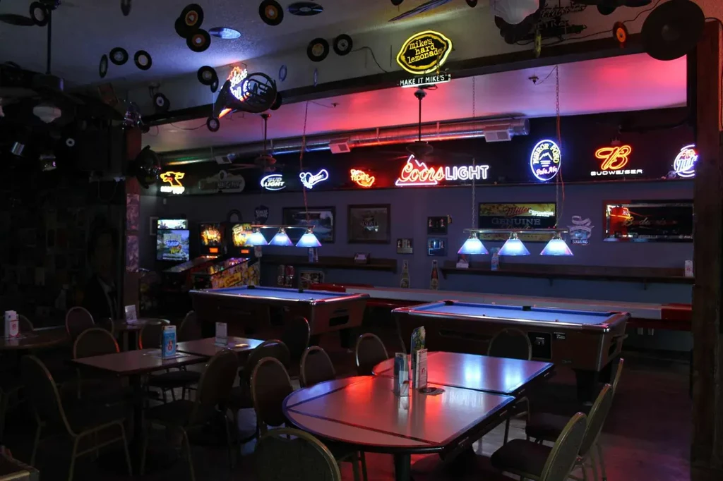 Dimly lit bar with pool tables, neon signs, vinyl records, arcade.