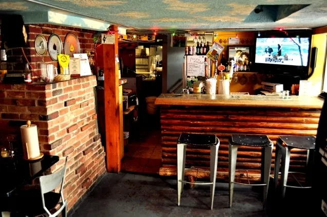 Rustic bar interior with wooden counter, metal stools, and TVs.