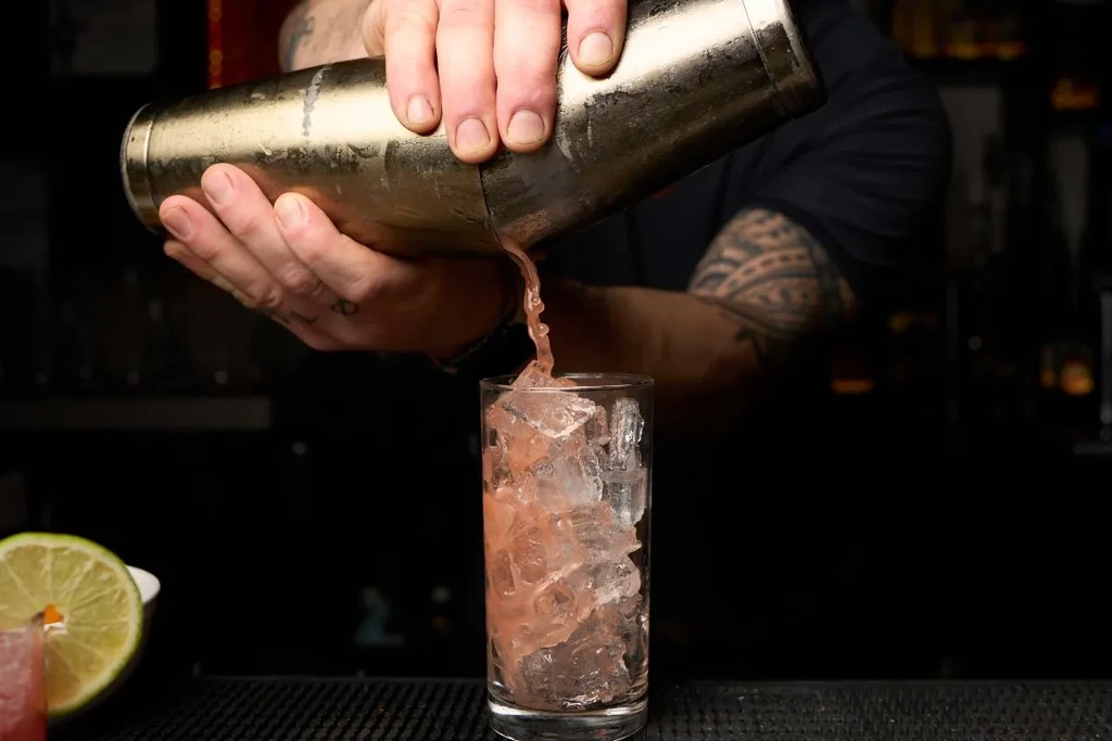 Tattooed hands pour a pink cocktail from shaker into an iced glass.