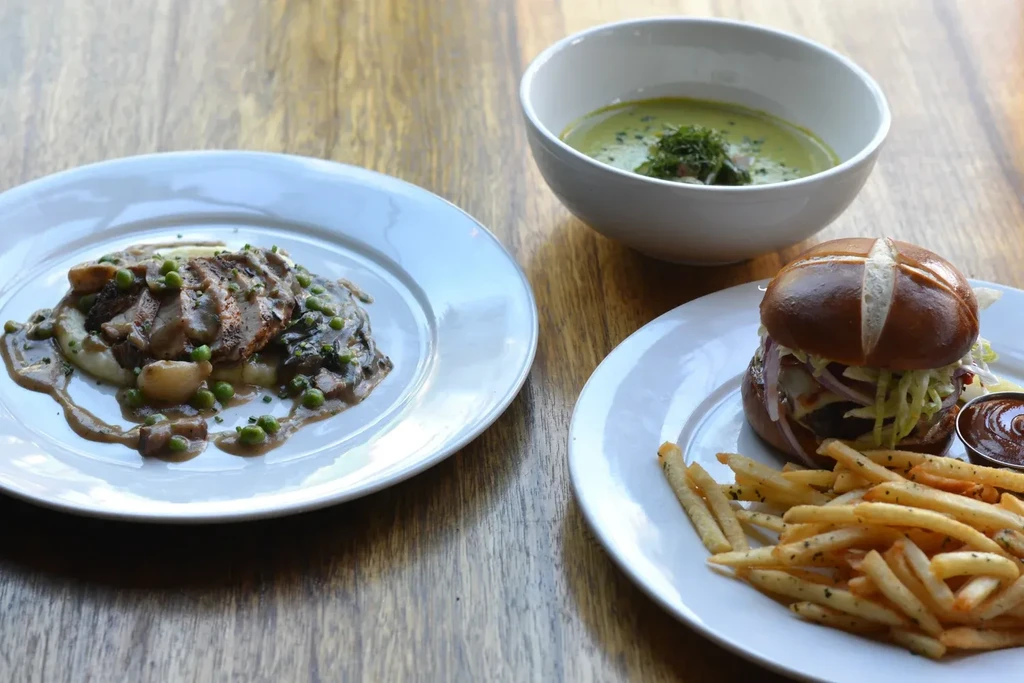 Grilled meat, pretzel burger, fries, and green soup on a wooden table.