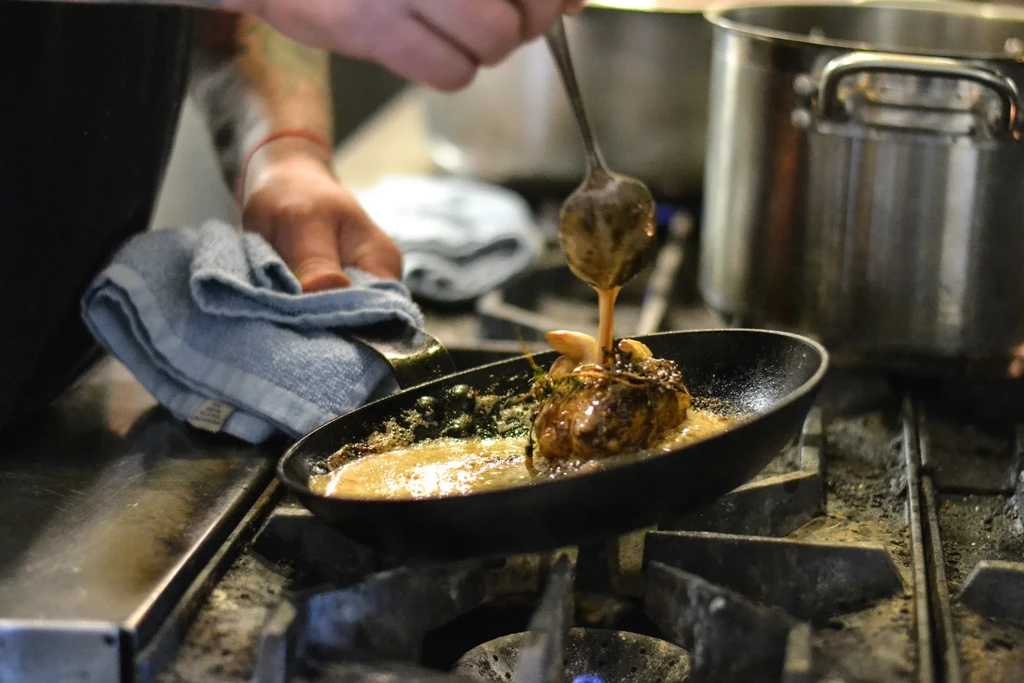 Chef's hands baste seared meat with herbs in a cast iron pan.