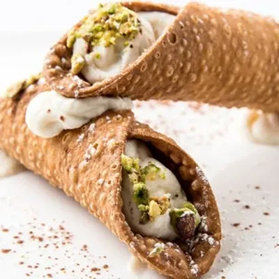 Two golden-brown cannoli with ricotta, pistachios, and cocoa specks.