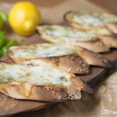 Cheesy bread with green herbs on a wooden board; lemon in background.