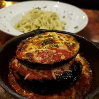 Skillet eggplant parmesan with melted cheese, herbs, and blurred pasta.