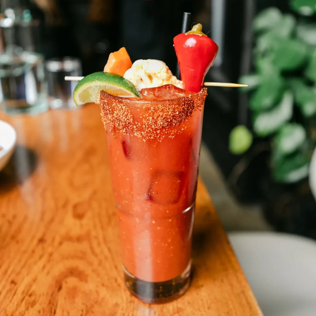 Spicy reddish-orange drink with skewered lime, cauliflower, and bell pepper.