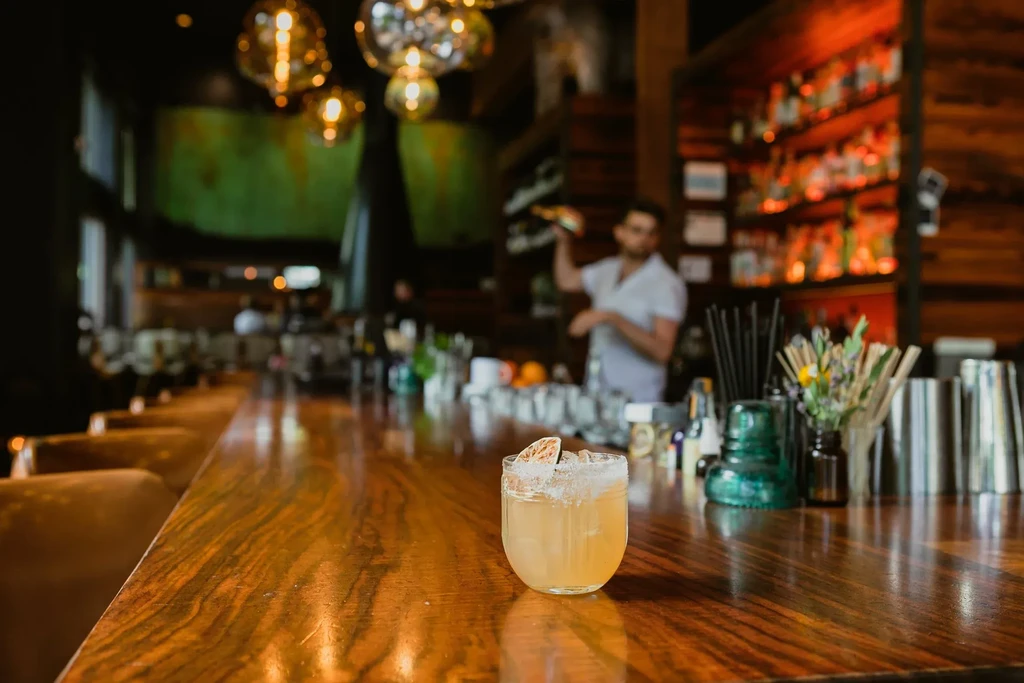 A cocktail with a salted rim on a wooden bar, blurred bartender behind.