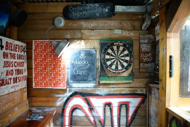 Heavily graffitied bar wall with dartboard, chalkboard, and a religious poster.